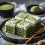 Four square pieces of matcha white chocolate mochi cake topped with a dusting of matcha powder and powdered sugar, served on a rustic black plate with a wooden spoon.